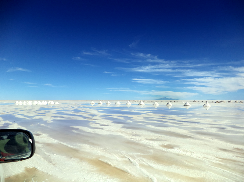 Super wide photograph of pale salt flats and vivid blue sky taken from the passenger seat of a vehicle. 